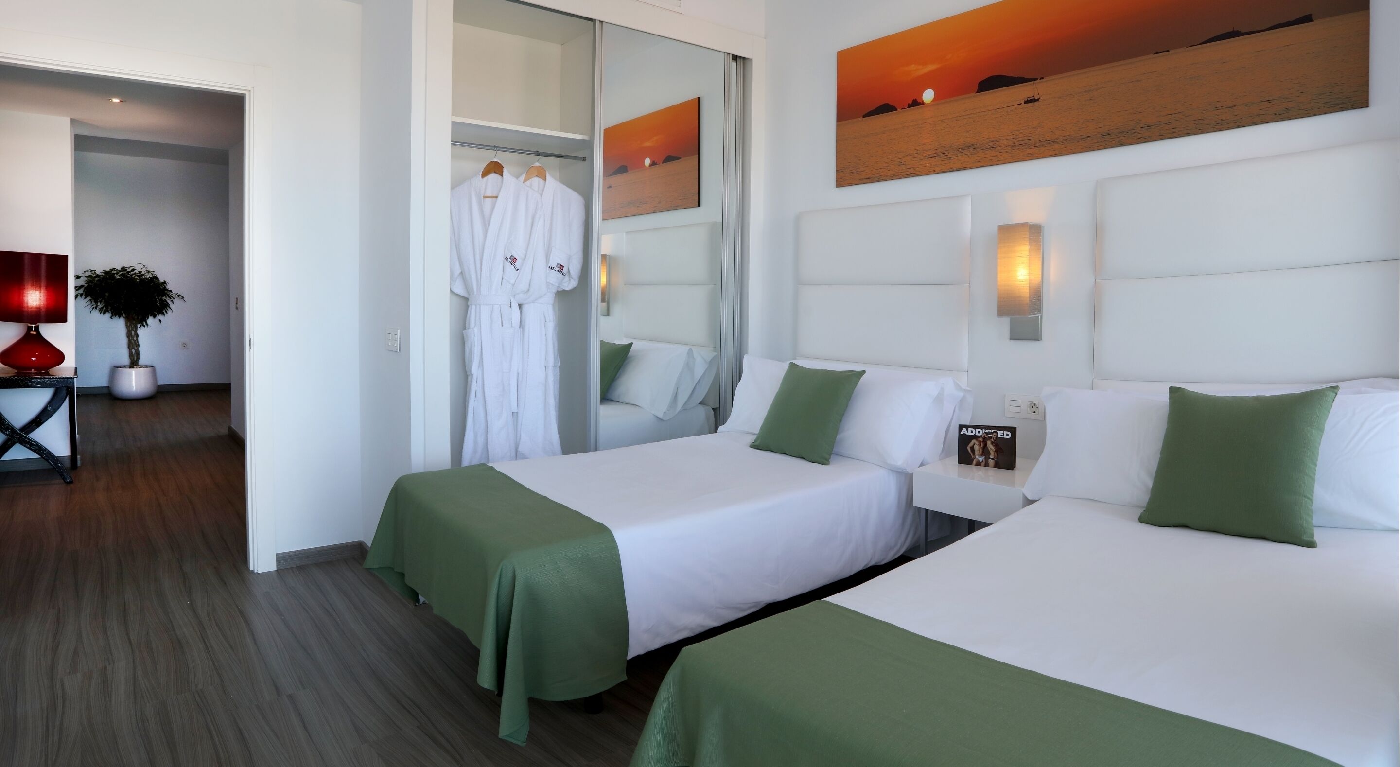 Habitación de hotel con dos camas, cubrecamas verdes, ropa de cama blanca, una mesita de noche con una lámpara, un cuadro de un atardecer y un espejo que refleja la habitación.