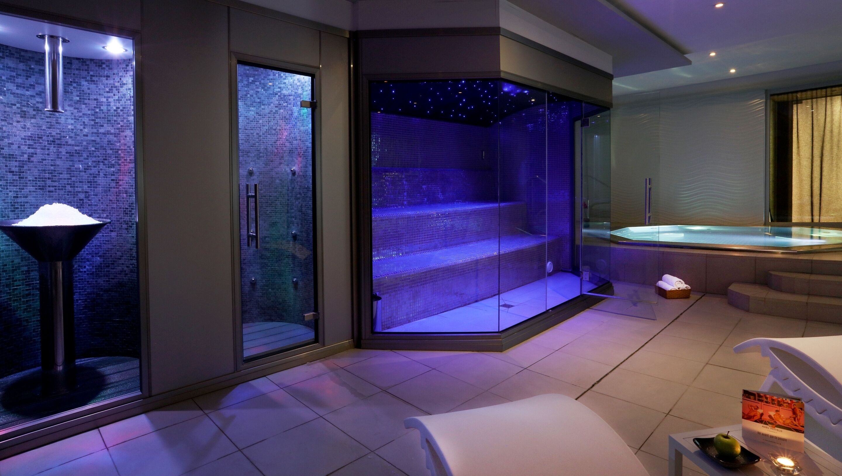 Een moderne spa-ruimte met een hottub in een glazen omhulsel, een massagestoel en een klein tafeltje met een kaars en een menukaart.