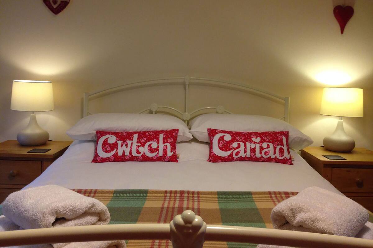 A neatly made bed with two pillows labeled 'Cwch' and 'Cariad', two bedside lamps, and a checkered blanket.