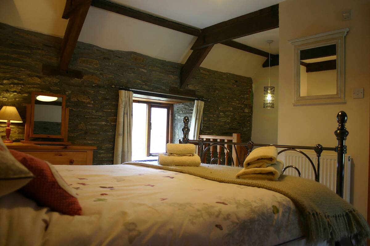 A bedroom with a stone wall, wooden beams, a bed with towels, a nightstand, a mirror, and a window with curtains.