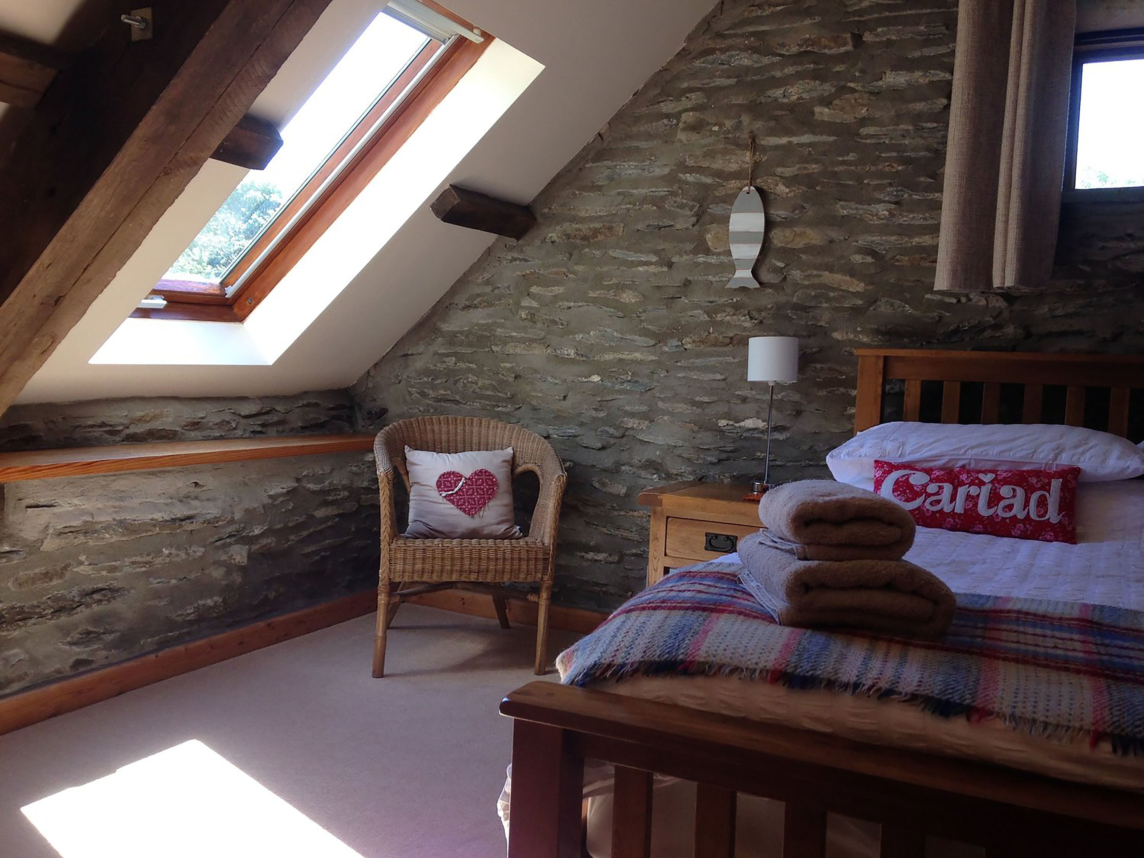 A cozy bedroom with a sloped ceiling, stone walls, a wicker chair, a bedside lamp, and a bed with a plaid blanket and a pillow with 'Cariad' written on it.