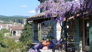 Un bâtiment en pierre avec un toit en tuiles, un balcon orné de plantes grimpantes aux fleurs violettes et un espace extérieur aménagé dans un style rustique.