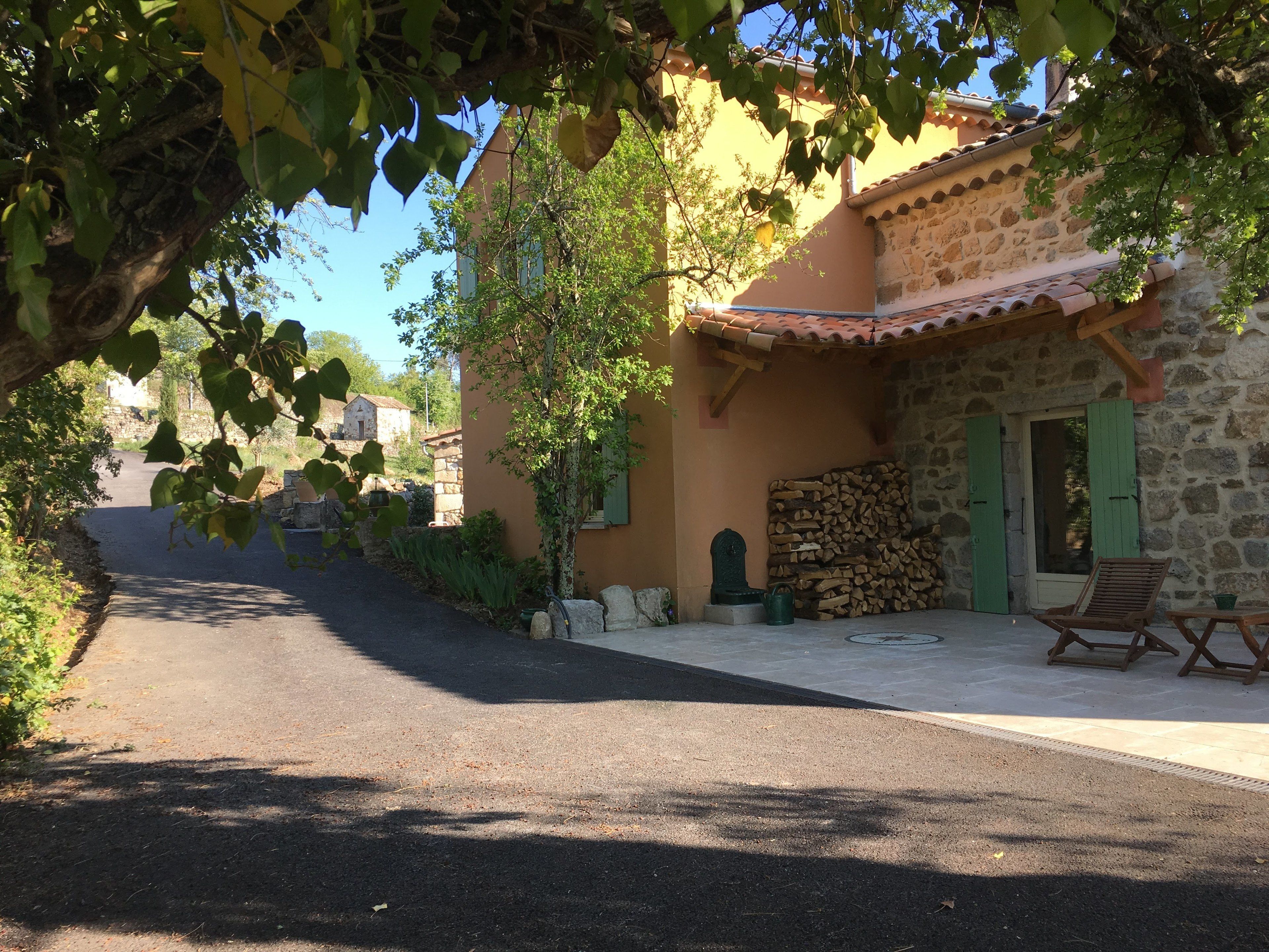 A narrow road leading to a stone building with a tiled roof, a stack of firewood, and a wooden chair outside.