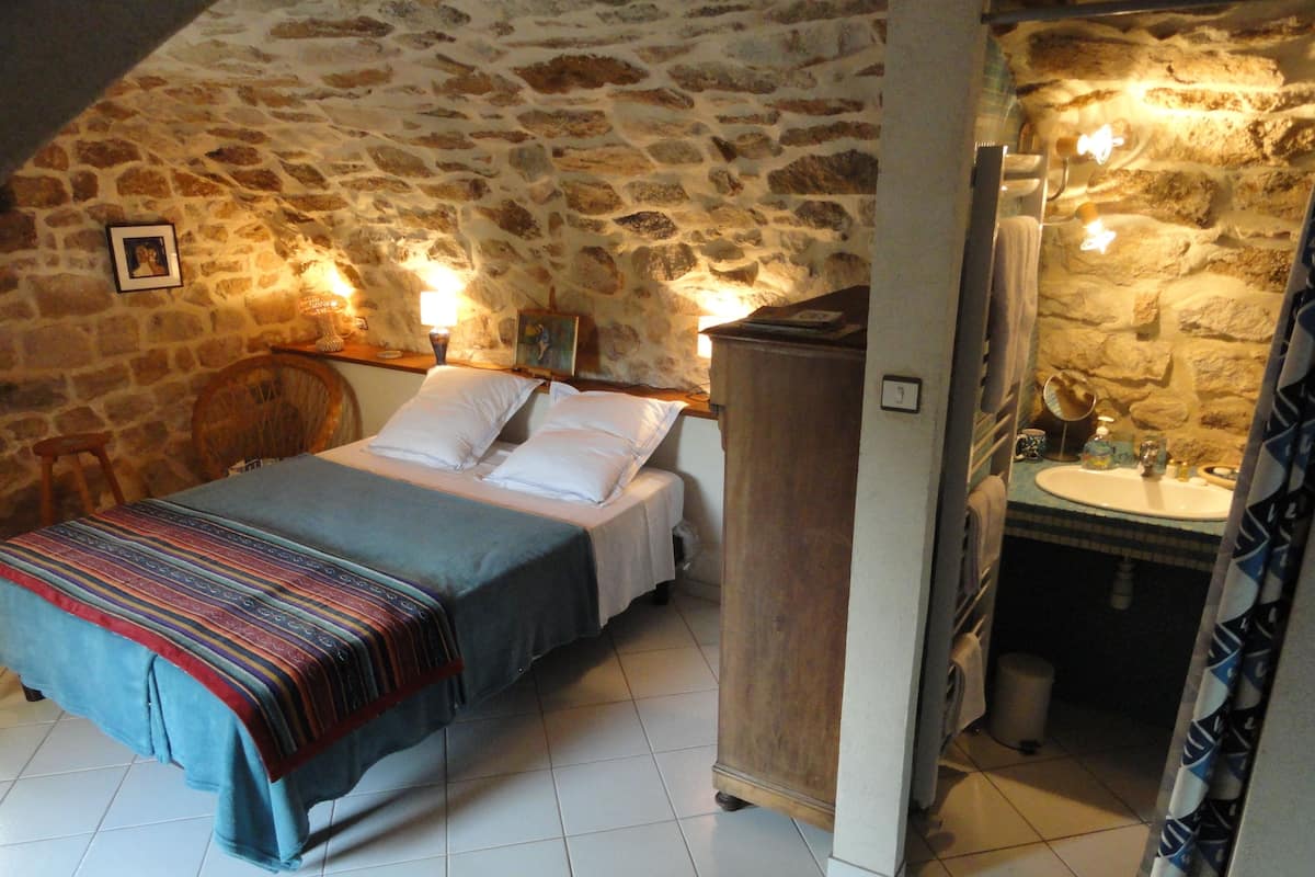 A bedroom with a stone wall, a bed with a colorful blanket, a nightstand, and a bedside table with a lamp.