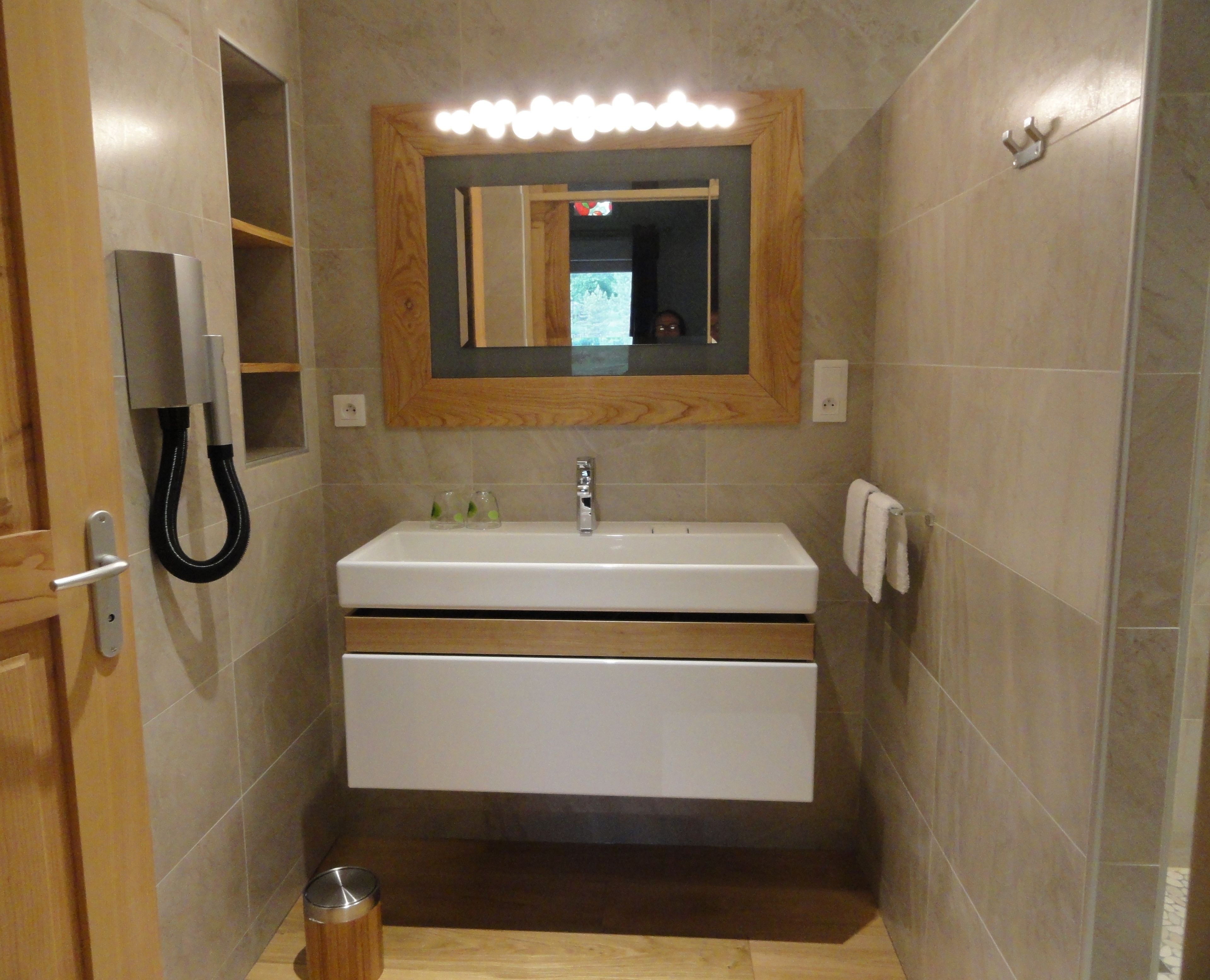 Un baño con un lavamanos blanco, un espejo con luces, una puerta de madera y un bote de basura.