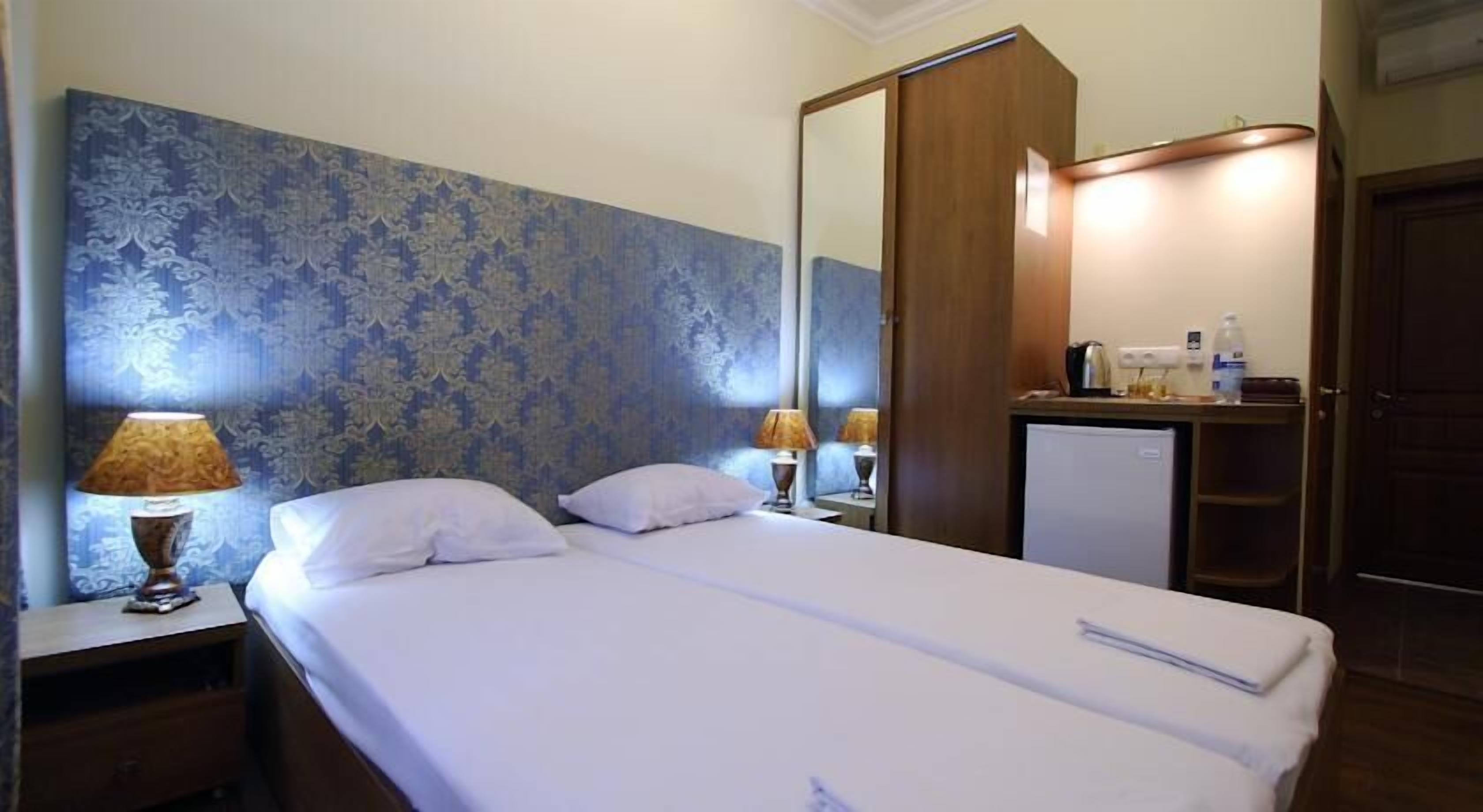 A hotel room with a bed, two bedside lamps, a mirror, a wardrobe, and a small desk with a refrigerator.