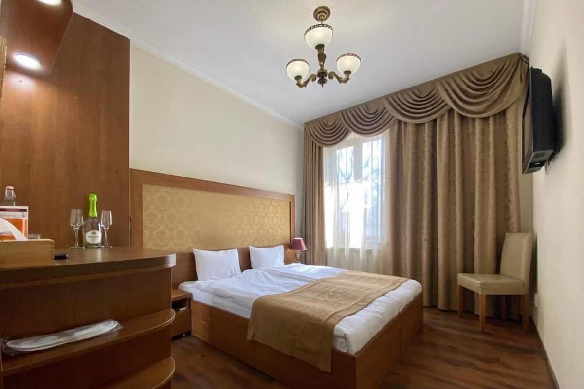 Habitación de hotel con cama de madera, escritorio con botella y vasos, una silla y televisión en la pared.