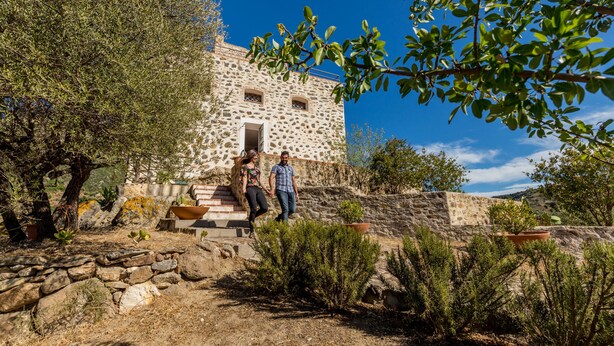 A stone building with a staircase leading to the entrance, surrounded by dry vegetation and a clear blue sky. - Castello Malicas (Galtelli)