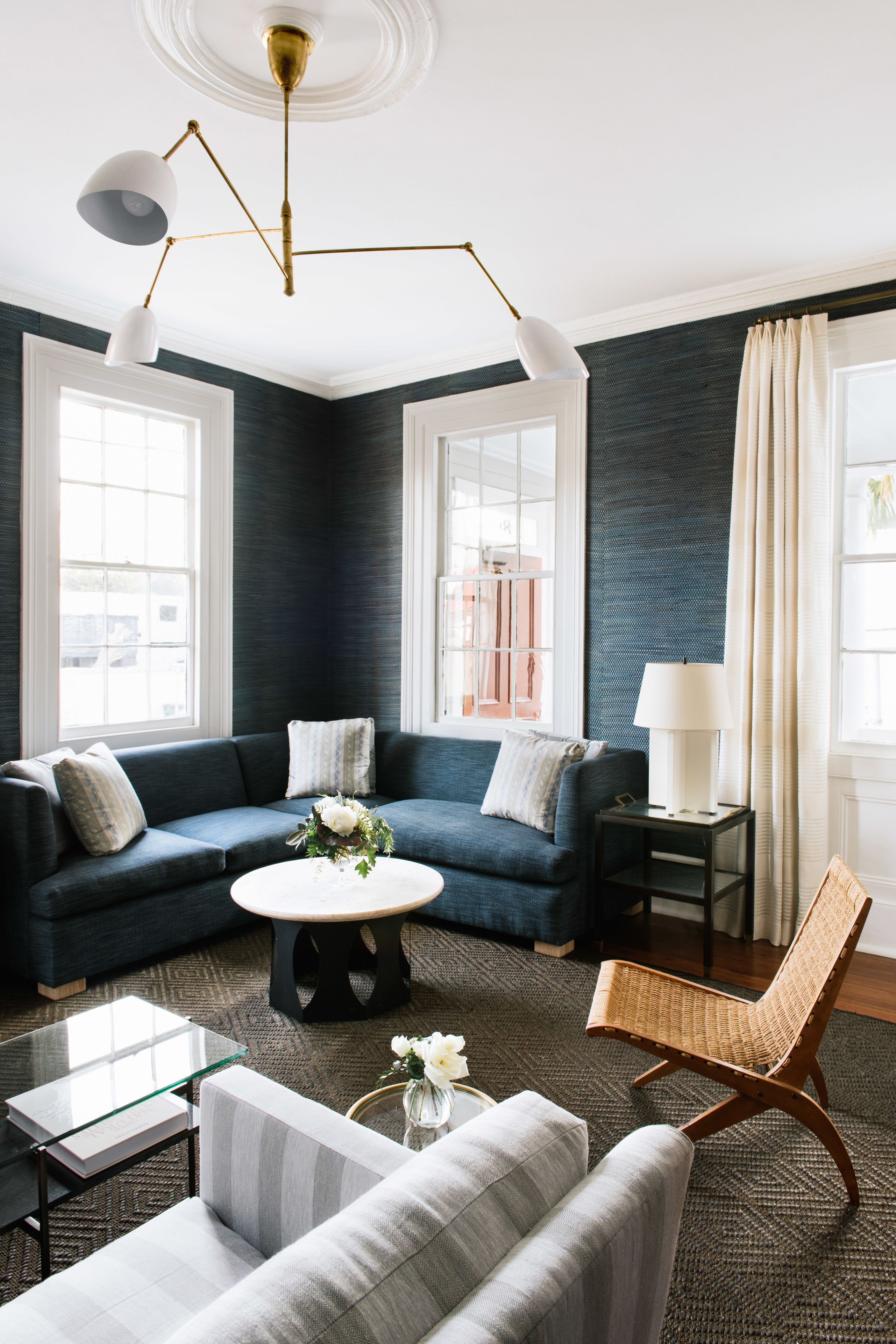 Ein modernes Wohnzimmer mit einer dunkelblauen Akzentwand, einem blauen Sofa, einem weißen Sessel, einem Glastisch und einem Korbstuhl.