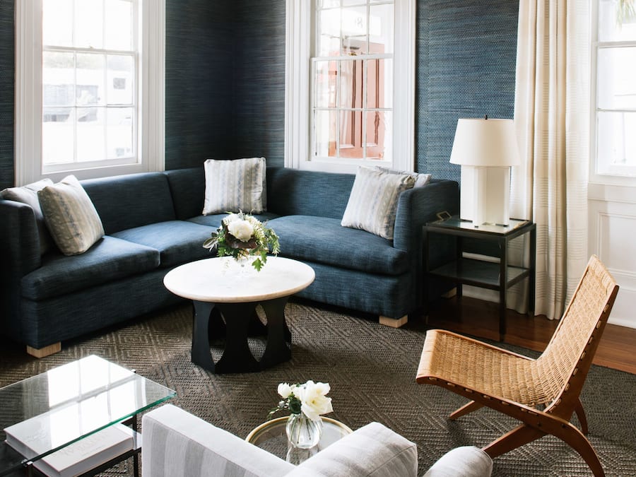 Ein modernes Wohnzimmer mit einer dunkelblauen Akzentwand, einem blauen Sofa, einem weißen Sessel, einem Glastisch und einem Korbstuhl.