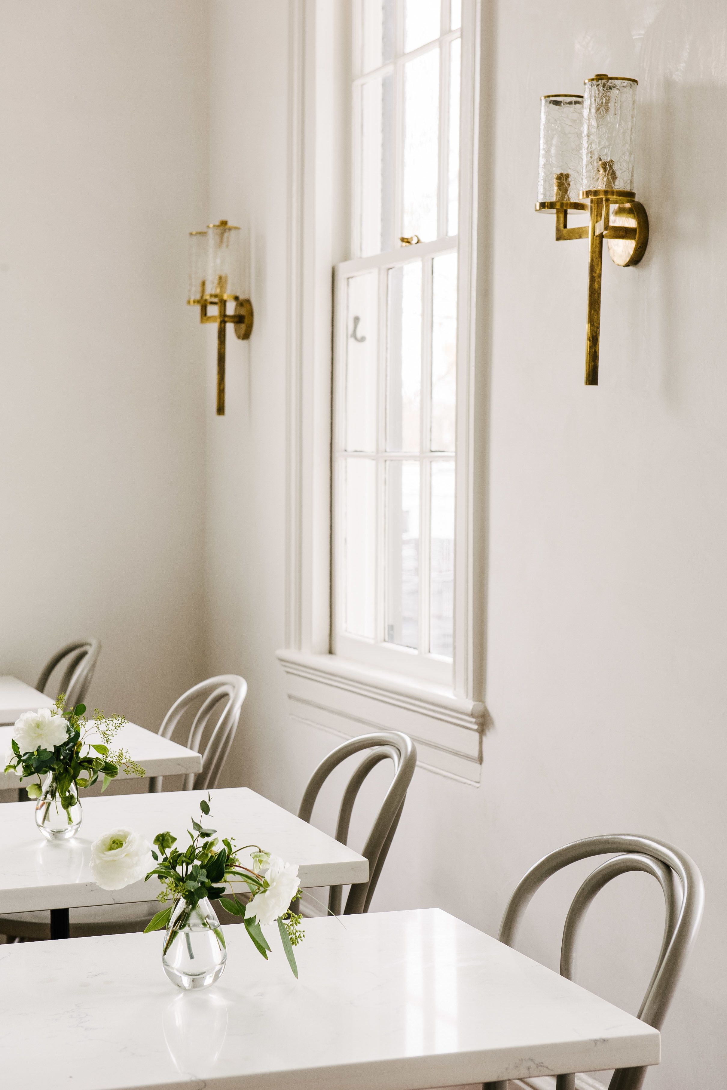 A dining area with white tables and chairs, a window with white frames, and wall-mounted light fixtures.