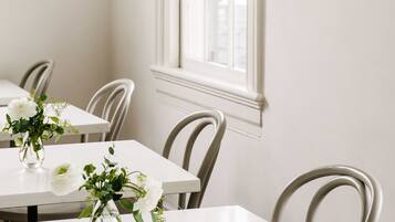 A dining area with white tables and chairs, a window with white frames, and wall-mounted light fixtures.
