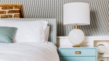 En pænt redt seng med hvide sengetøj og en teal farvet pude, en hvid lampe på et blåt natbord, og tapet med et mønster i baggrunden.