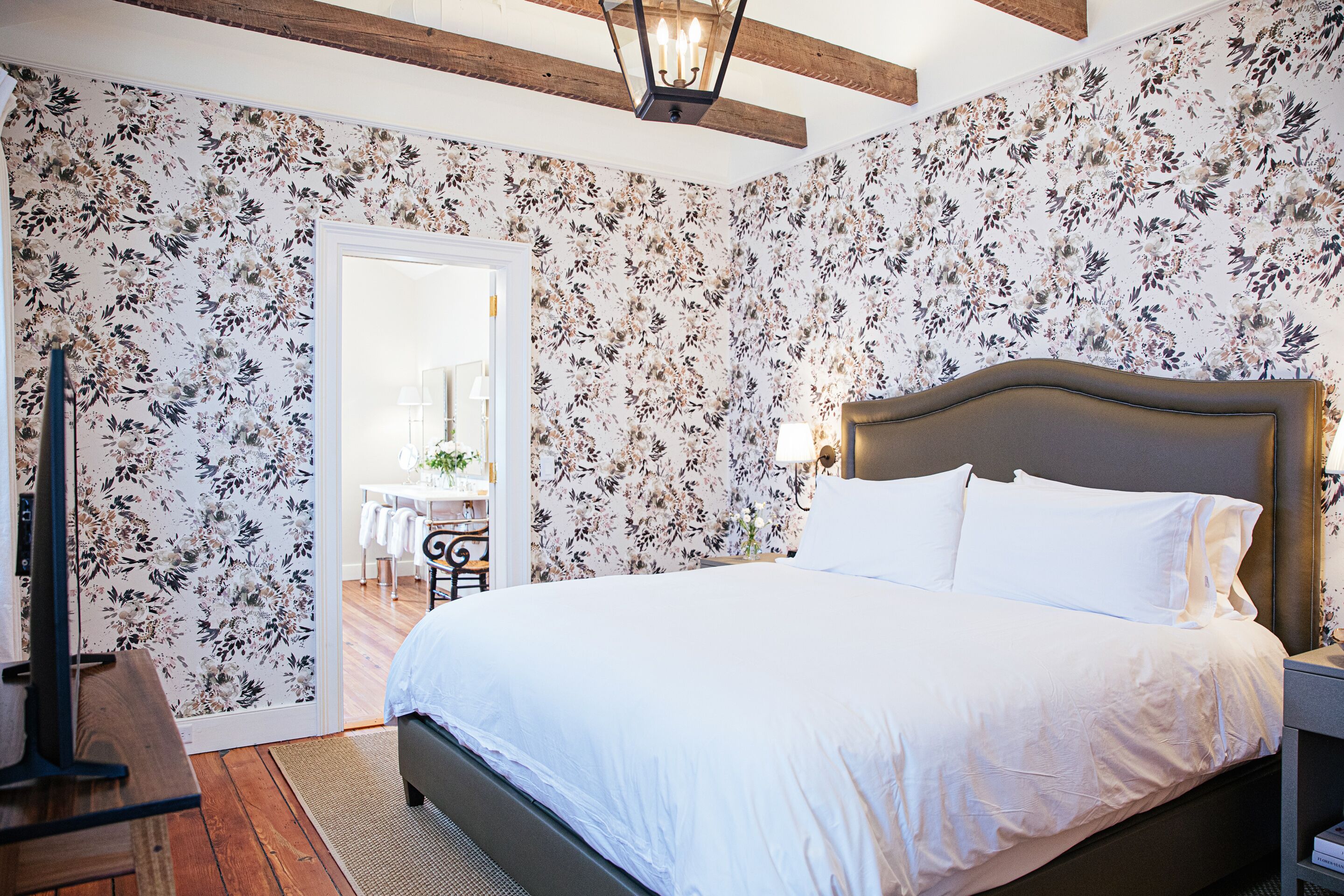 Un dormitorio con una cama grande, papel tapiz floral, suelo de madera y una mesita de noche con una lámpara.