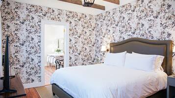 Un dormitorio con una cama grande, papel tapiz floral, suelo de madera y una mesita de noche con una lámpara.
