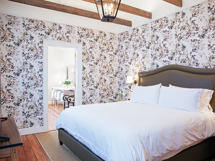 A bedroom with a large bed, floral wallpaper, a wooden floor, and a nightstand with a lamp.