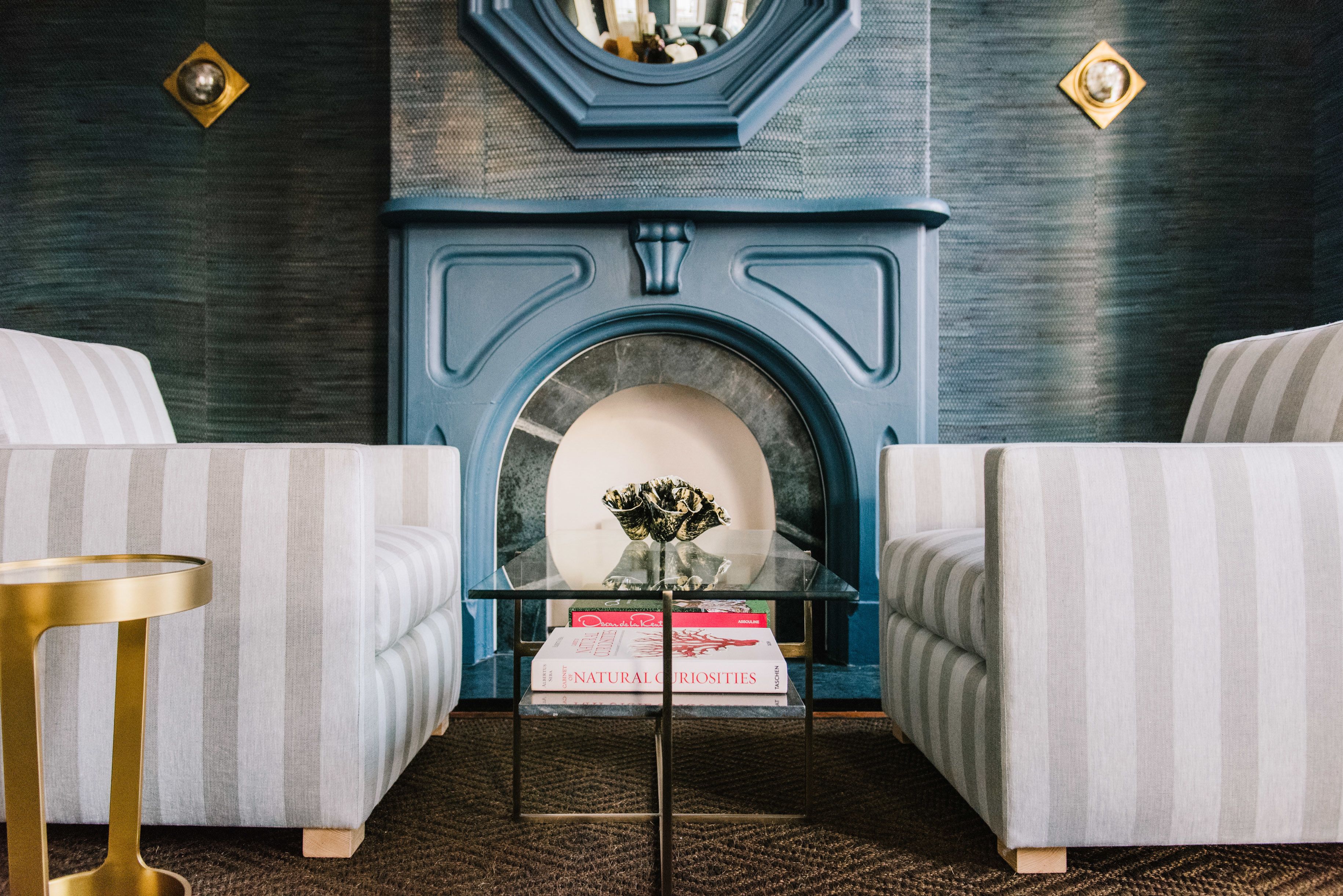 A room with two striped armchairs, a glass coffee table with books, and a blue fireplace.