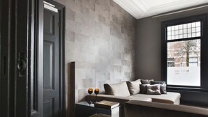Una sala de estar moderna con pared de azulejos grises, un sofá con cojines, una mesa de centro y una mesita de noche con una lámpara y libros.