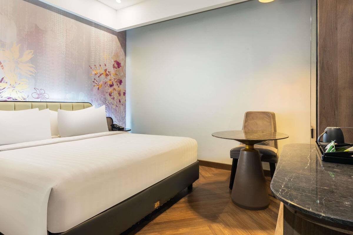 A hotel room with a bed, a small round table with a chair, a desk with a chair, and a large floral artwork on the wall.