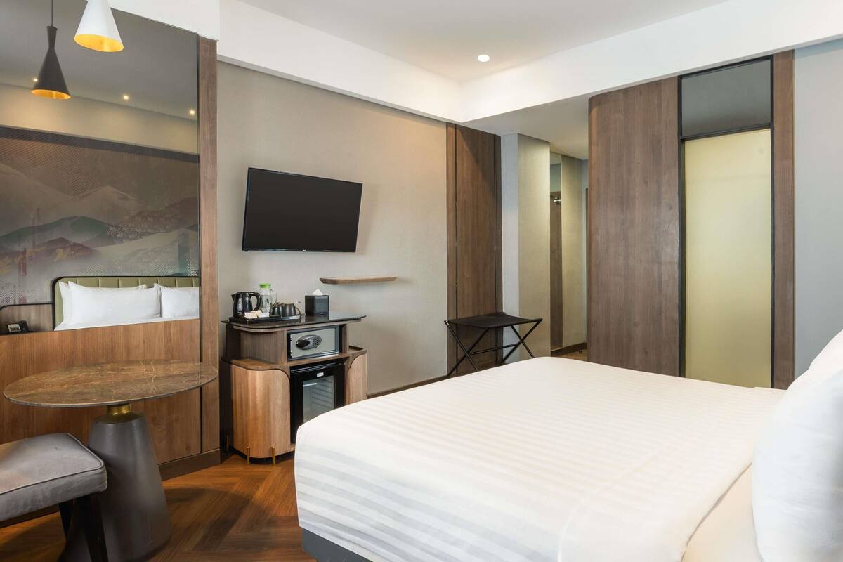 A modern hotel room with a bed, a desk, a chair, a television, and a small table with a kettle and cups.