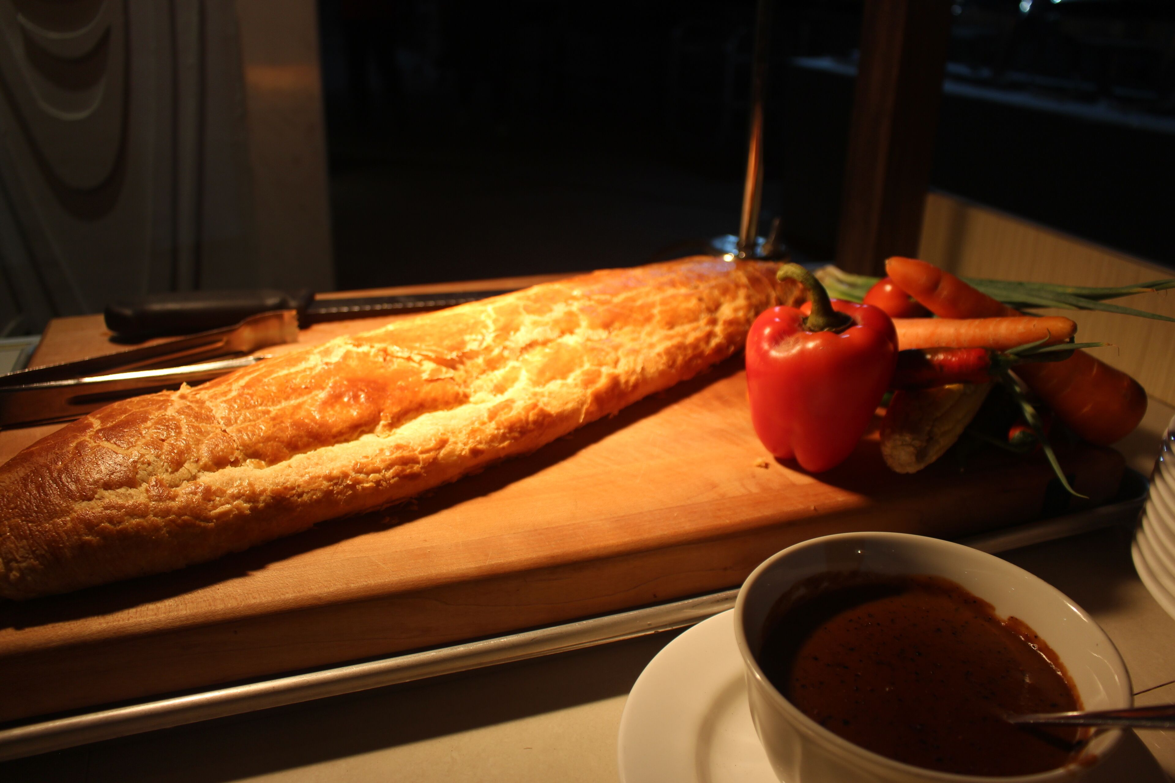 En lång baguette på en träskärbräda, en skål med soppa och några grönsaker, inklusive morötter och tomater.