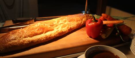 Una baguette larga sobre una tabla de madera, un tazón de sopa y algunas verduras, incluyendo zanahorias y tomates.