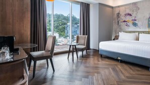 Ein Hotelzimmer mit einem großen Bett, zwei Sesseln, einem kleinen Tisch und Blick auf eine Stadtlandschaft durch große Fenster.