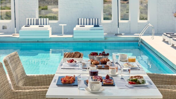 Ein Frühstückstisch mit Korbstühlen am Pool, mit Blick auf einen Pool, eine weiße Wand und Fenster.