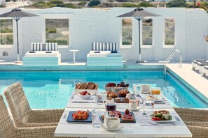 A poolside breakfast setup with wicker chairs and a table, overlooking a pool with a white wall and windows.