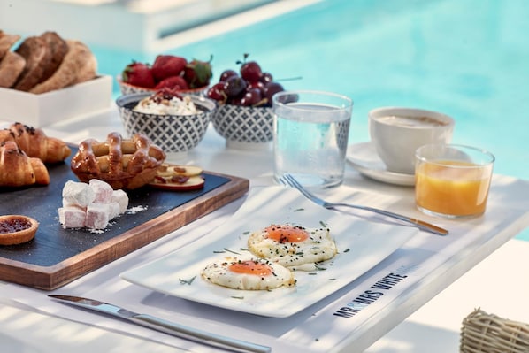 A breakfast setting with croissants, Turkish delight, and a plate of poached eggs, alongside a glass of orange juice and a cup of coffee by a pool.