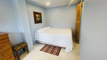 A bedroom with a bed, a wooden dresser, a bench, a painting on the wall, and a door.