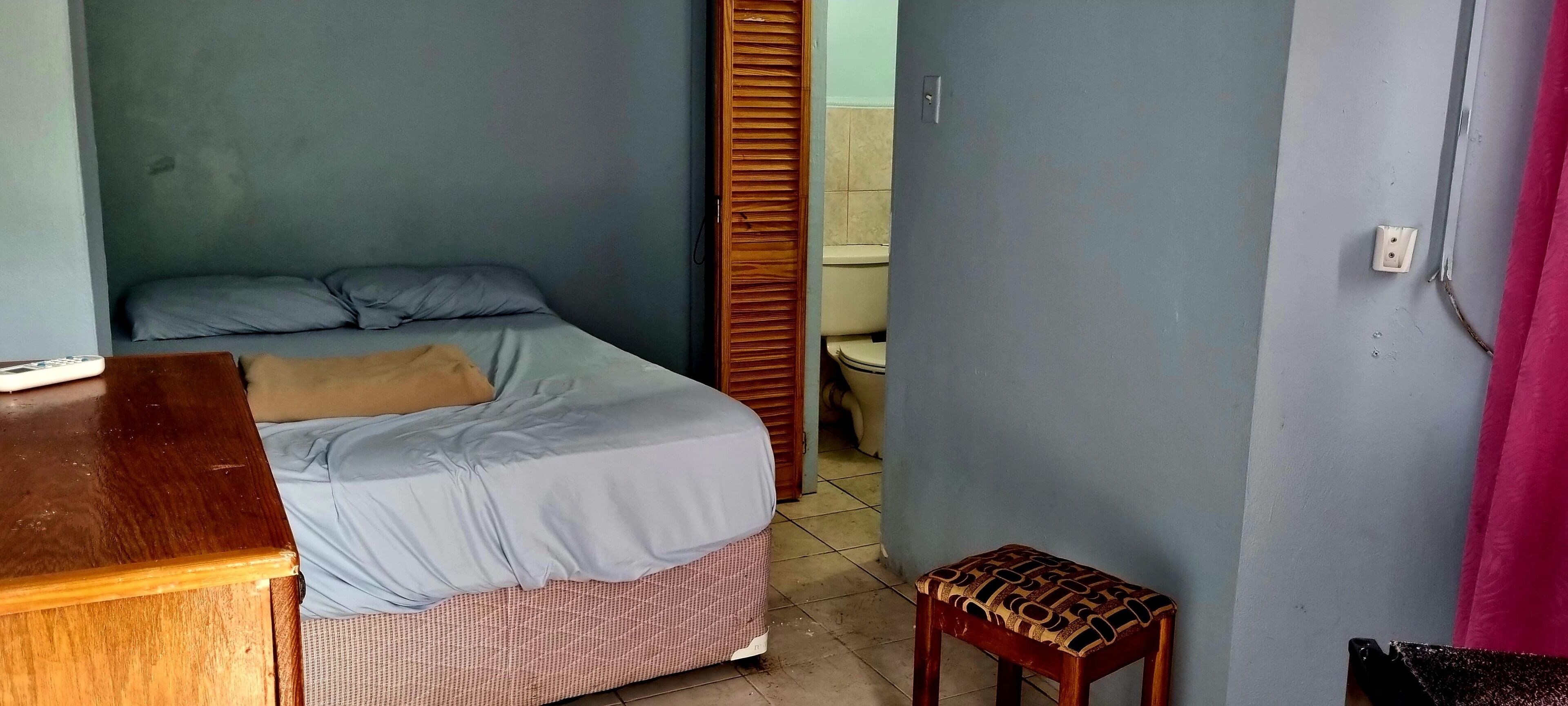 Una habitación pequeña y funcional con una cama, un escritorio de madera, un taburete y un inodoro.