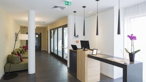 A modern reception desk with a computer, a potted plant, and a view of a hallway with a sofa and lamp. - Livris Hotel (Zagreb)