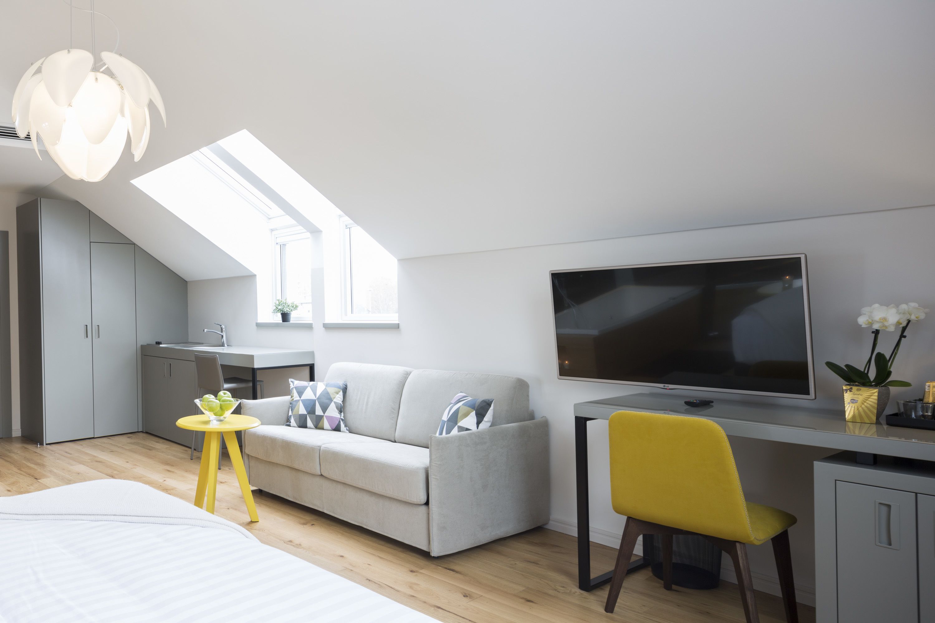A modern, compact living space with a loft bedroom, a kitchenette, a grey sofa, a yellow chair, and a large TV.