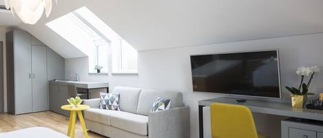 A modern, compact living space with a loft bedroom, a kitchenette, a grey sofa, a yellow chair, and a large TV.
