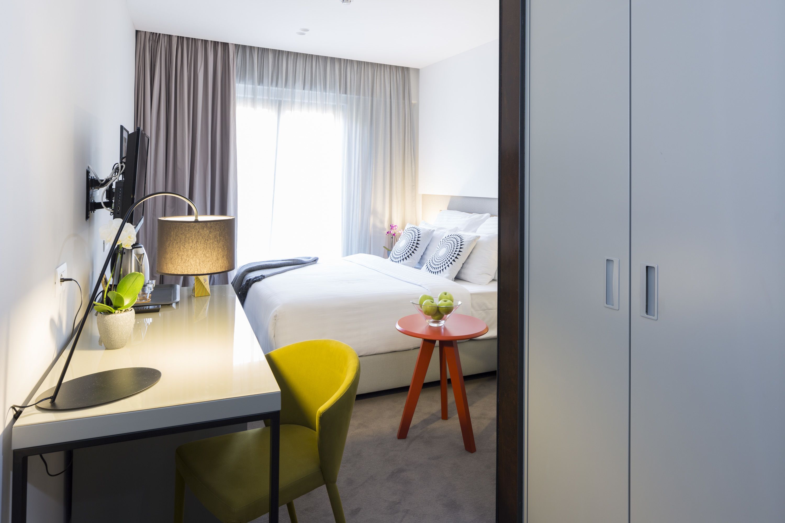 Une chambre d’hôtel avec un lit, un bureau avec une lampe, une chaise, une petite table avec un bol de fruits et un téléviseur fixé au mur.