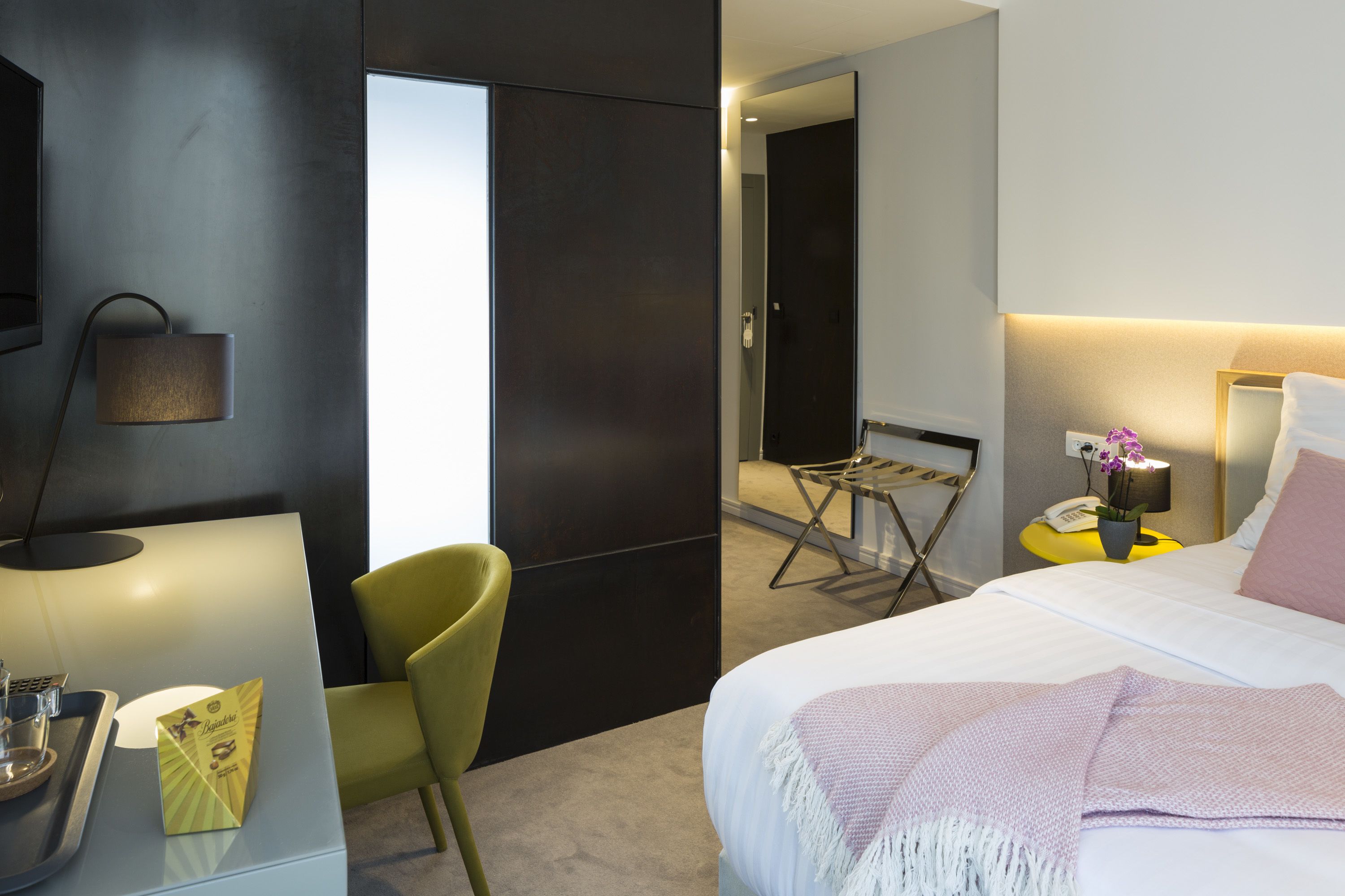 A modern hotel room with a bed, bedside table, lamp, chair, and a view of the hallway.