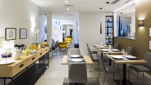 A modern dining area with wooden tables and chairs, a long counter with a coffee station, and a bright interior with natural light. - Livris Hotel (Zagreb)