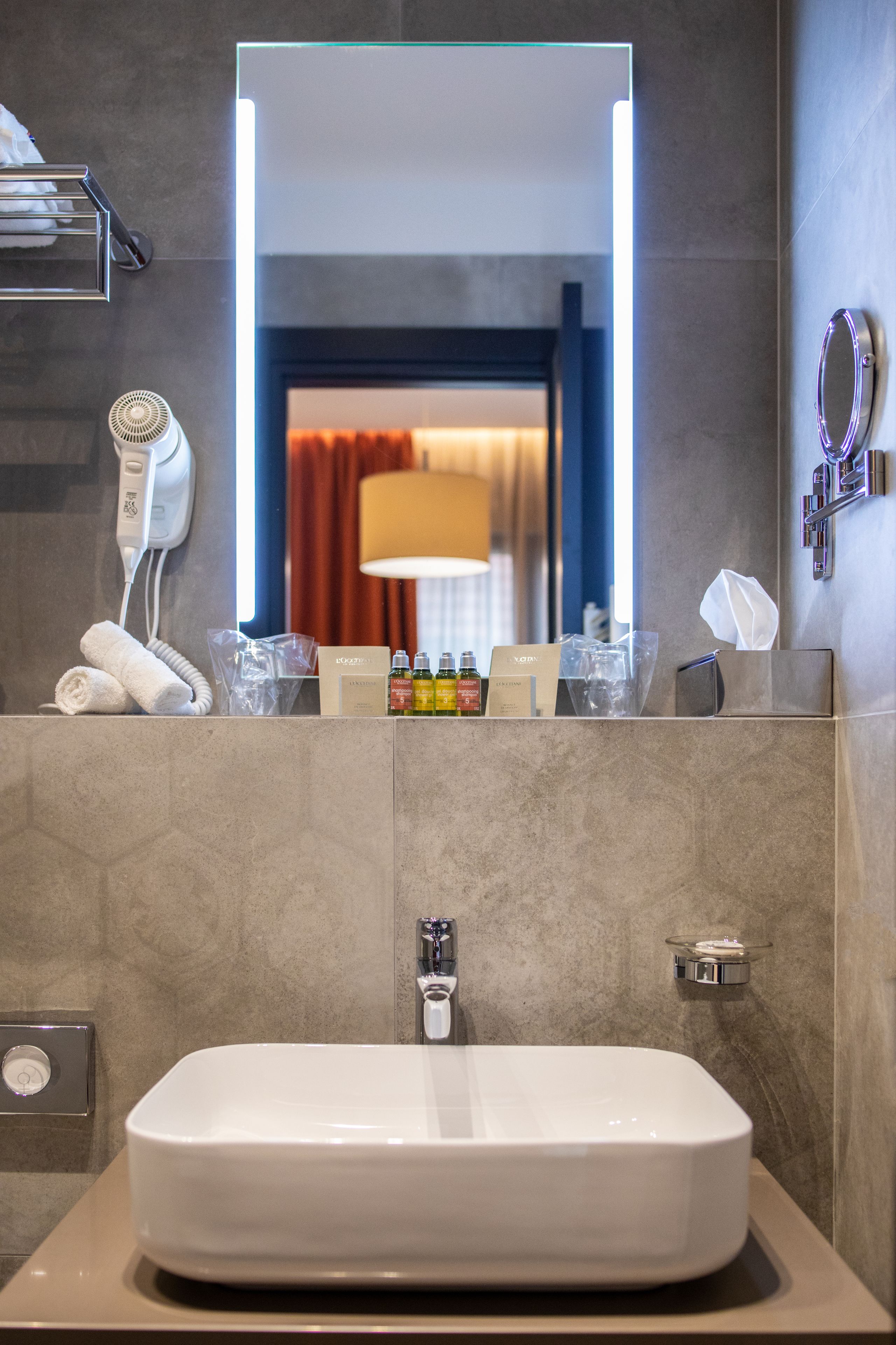 A modern bathroom with a large mirror, a white sink, a hairdryer, toiletries, and a towel rack.