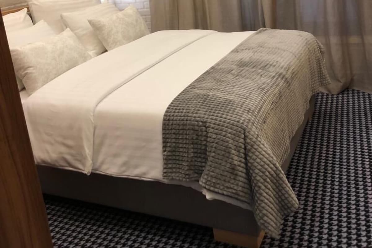 A hotel room with a brick-patterned wall, a bed with white bedding, a grey blanket, and a patterned carpet.