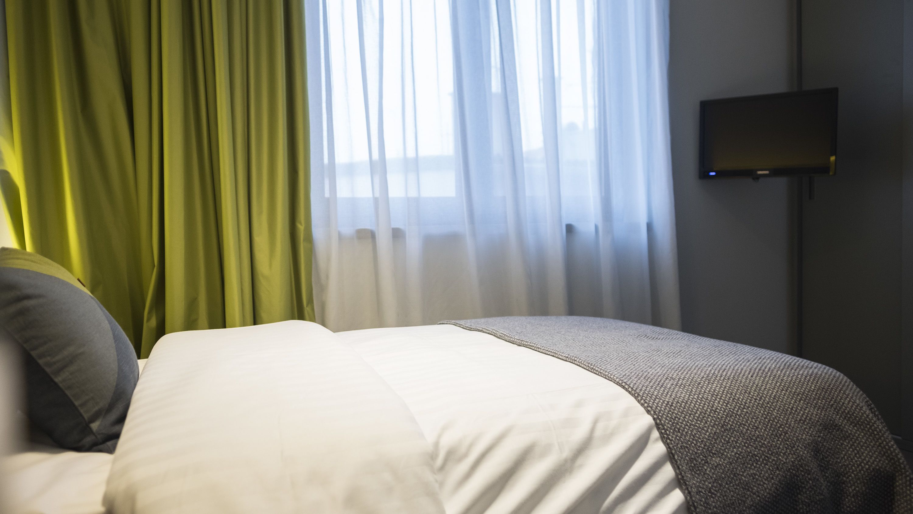 Une chambre d’hôtel avec un lit, des rideaux verts, une couvre-lit blanche et un téléviseur à écran plat fixé au mur.