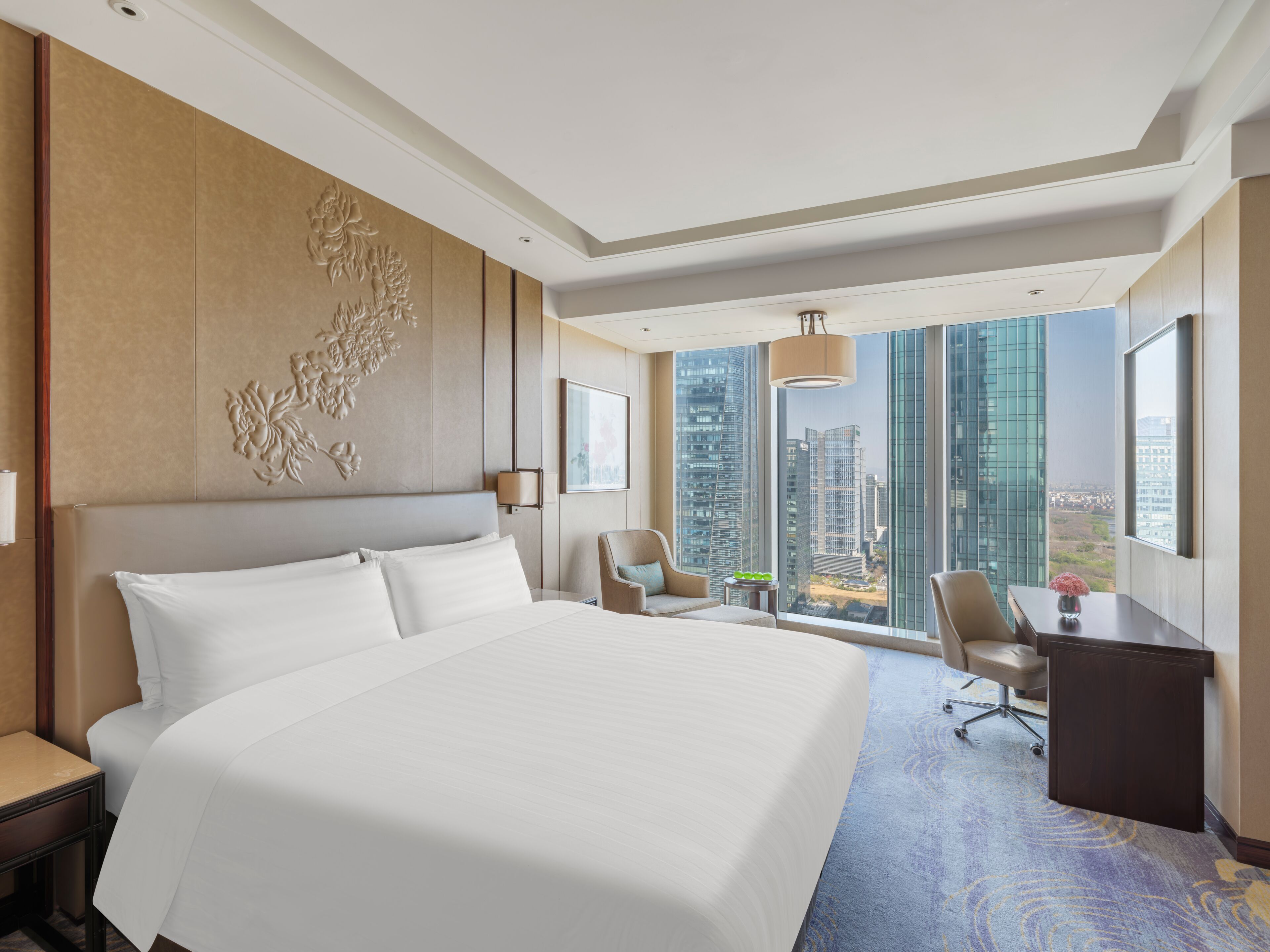 Una habitación de hotel con una cama grande, un escritorio, dos sillas, vistas a edificios altos y un panel decorativo en la pared.