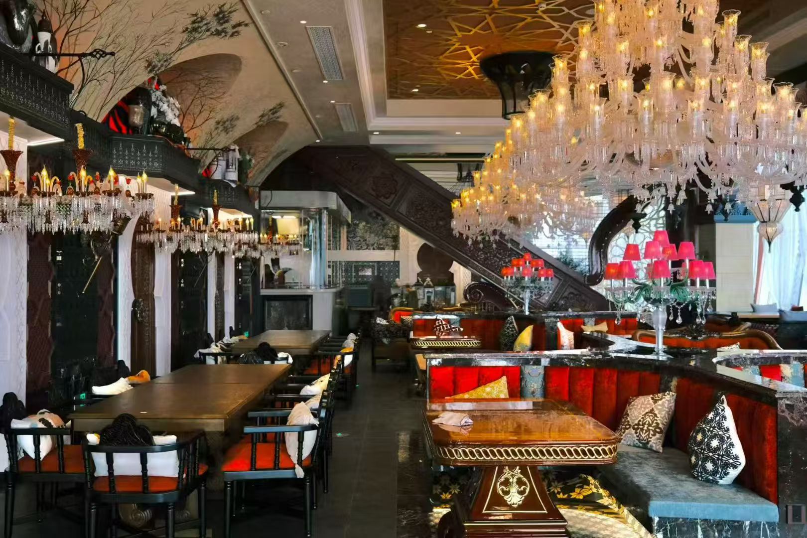 A restaurant interior with multiple dining tables and chairs, a grand chandelier, and ornate wall decorations.