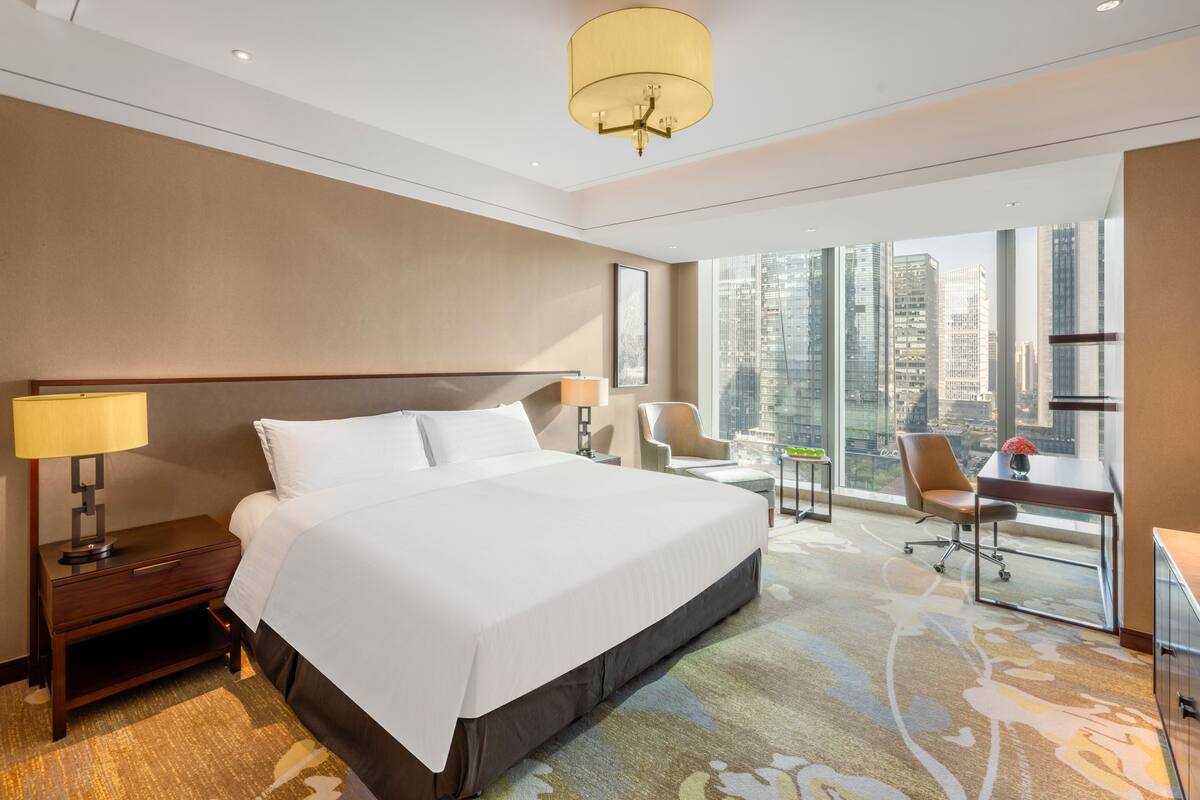 Habitación de hotel moderna con una cama grande, un escritorio, una silla y vistas a la ciudad a través de amplios ventanales.