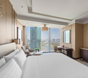 Een moderne hotelkamer met een groot bed, een bureau, een stoel en uitzicht op een stadsbeeld door grote ramen.