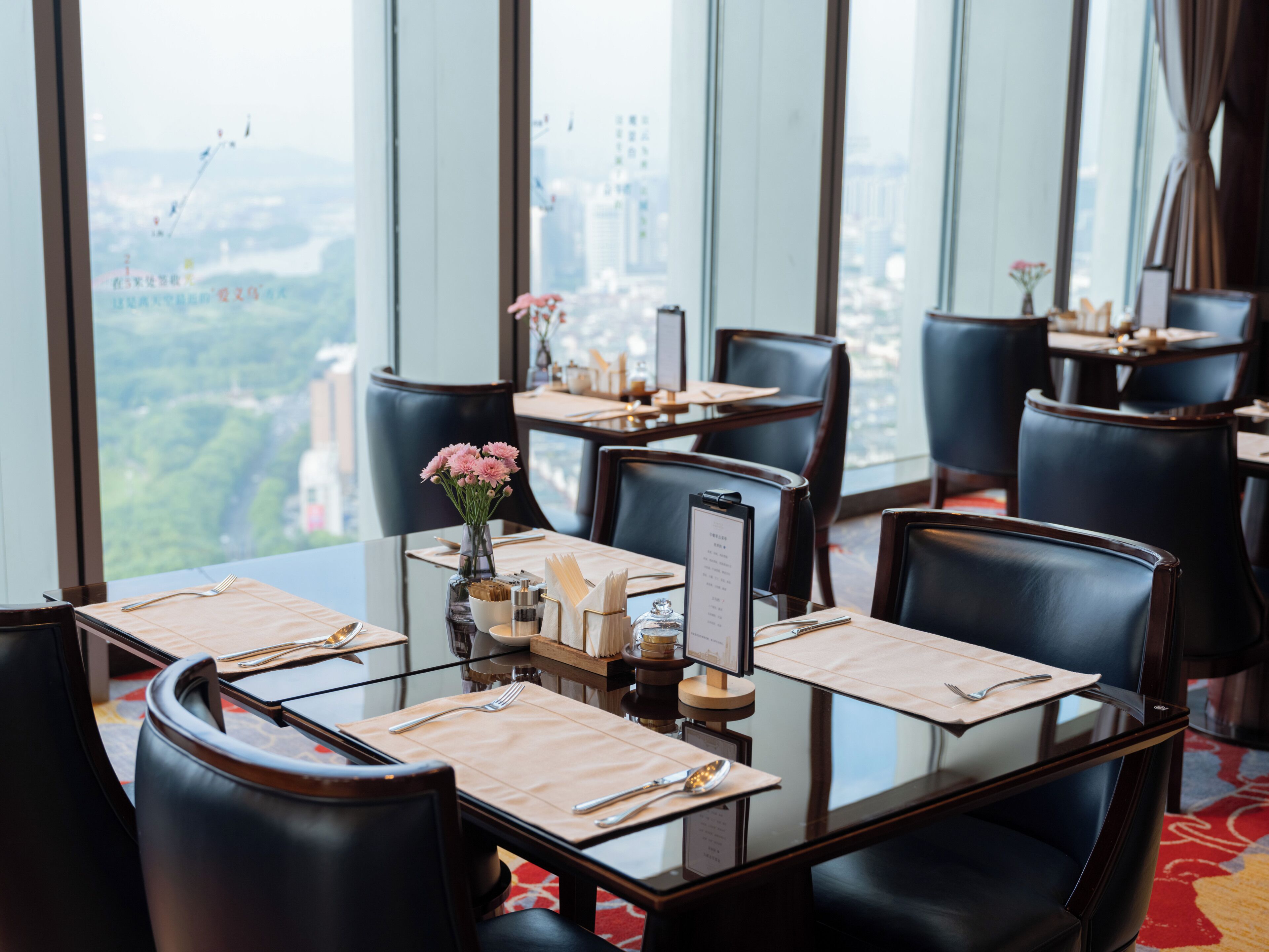 A restaurant with a city view, tables set with cutlery, and a vase with flowers.