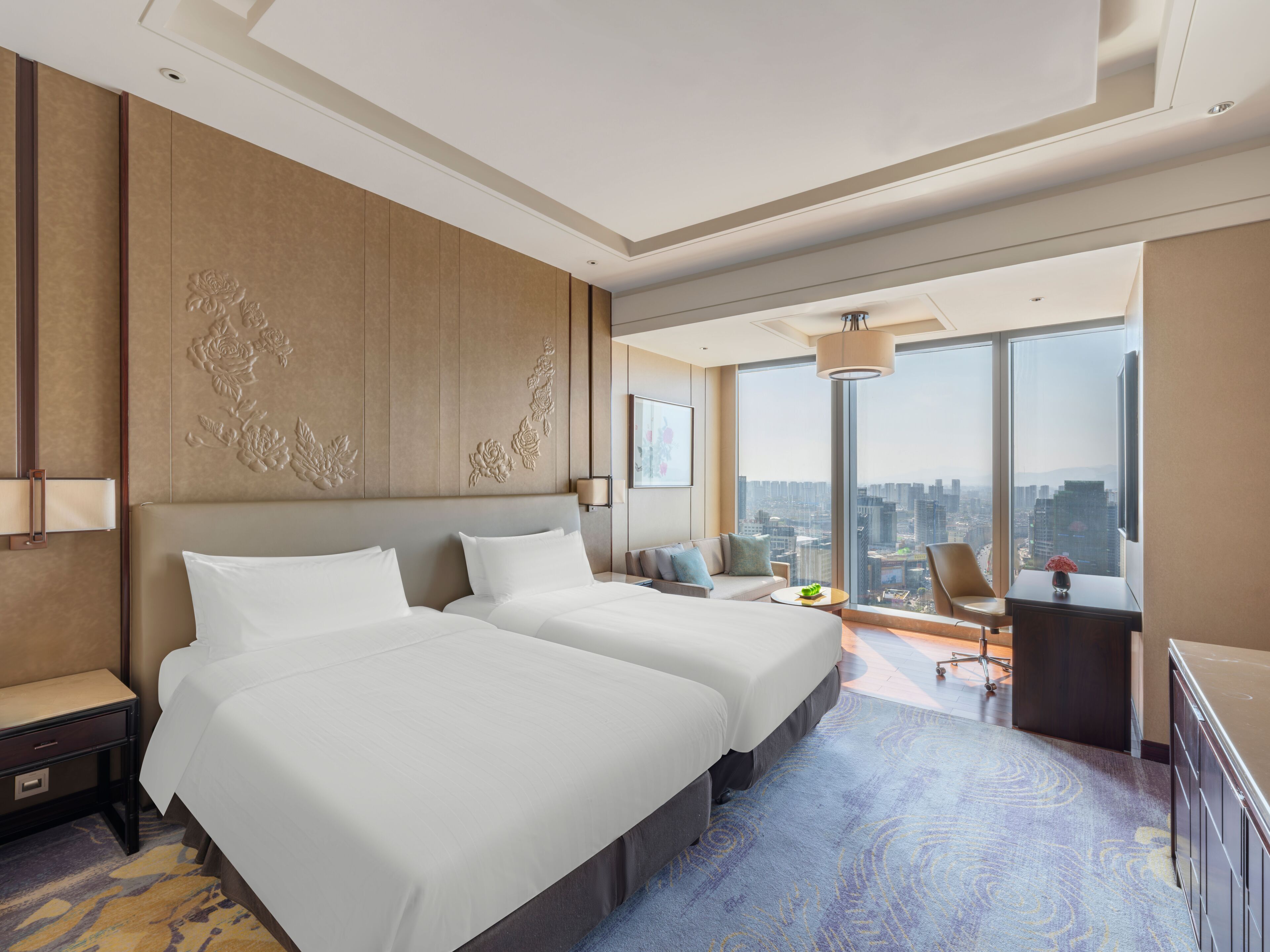 A hotel room with two beds, a desk, a chair, a large window with a city view, and a decorative wall panel.