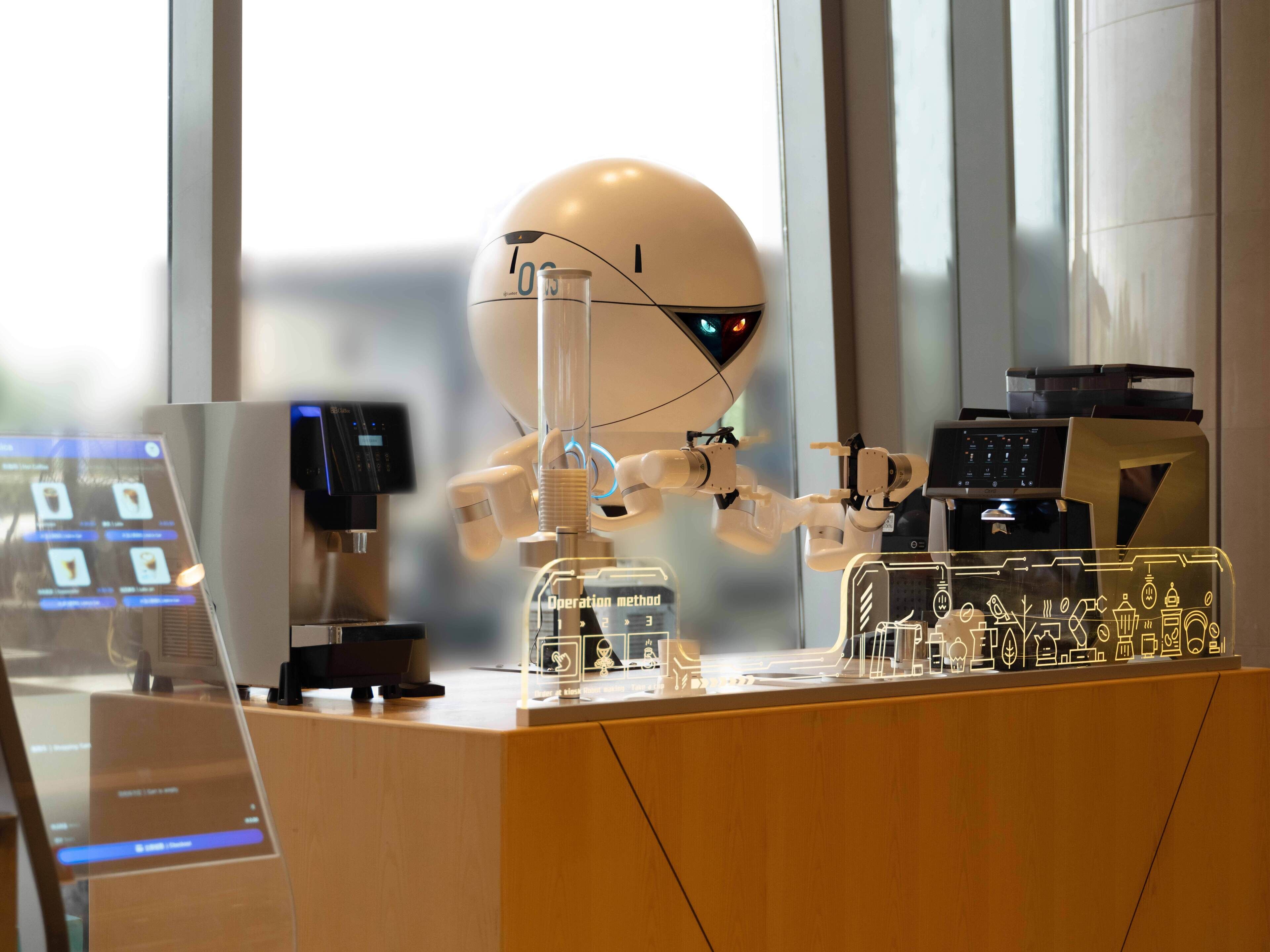 A robot interacting with a coffee machine and a computer monitor in a modern office setting.
