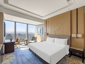 Una habitación de hotel moderna con una cama grande, un escritorio, una silla, una mesita con flores y un ventanal con vistas a la ciudad.
