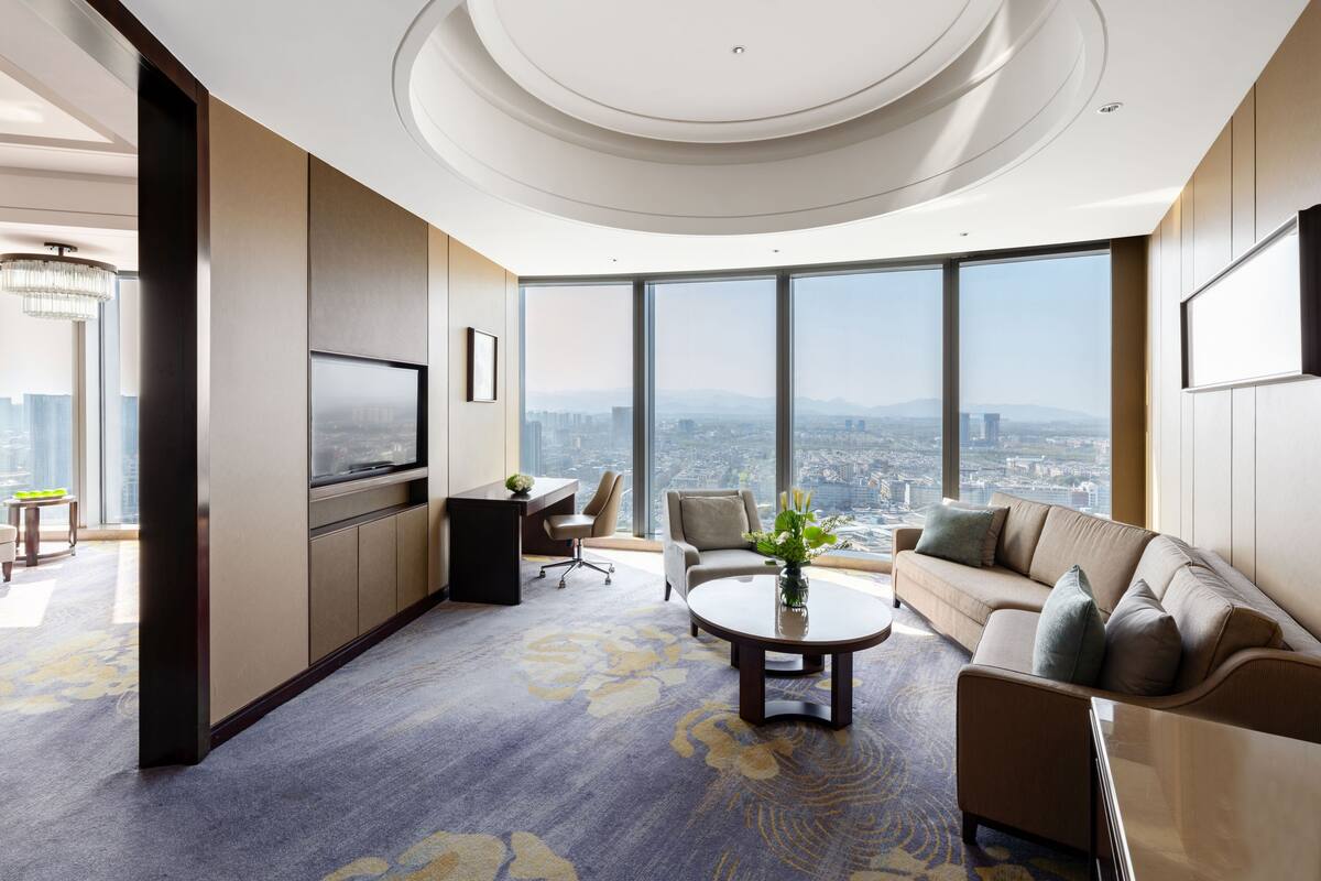 Habitación de hotel moderna con un ventanal grande que ofrece vistas a la ciudad, una mesa de centro redonda y una zona de estar acogedora.